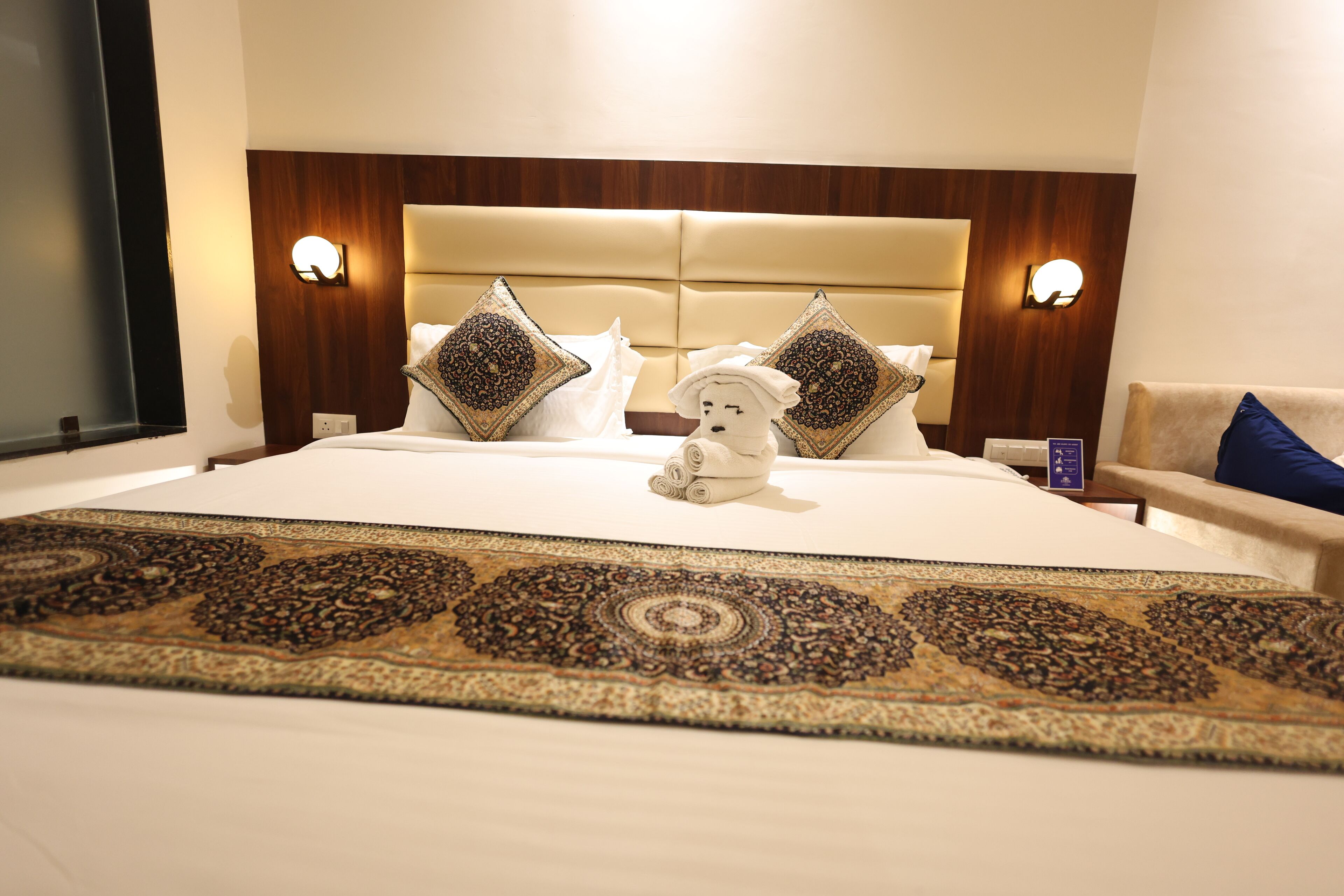 Ein Hotelzimmer mit einem großen Bett, einem Kopfteil mit integrierter Beleuchtung, dekorativen Kissen, einem Kuscheltier und einem gemusterten Teppich.