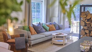 A modern living room with a grey sectional sofa, a round coffee table, and a view of a garden through large windows. - Holiday home in Liereman nature reserve (Oud-Turnhout)