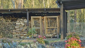 A rustic cabin with a stone wall and wooden doors, surrounded by a garden with various plants and a pile of firewood. - Holiday home in Liereman nature reserve (Oud-Turnhout)