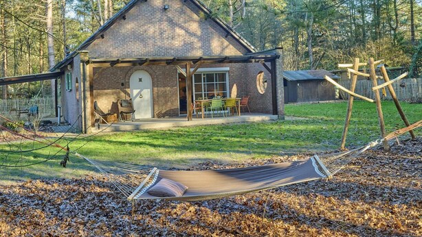 A rustic cabin with a covered porch, a hammock, and a wooden swing set in a wooded area. - Holiday home in Liereman nature reserve (Oud-Turnhout)