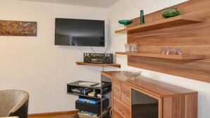 Een woonkamer met een flatscreen televisie, een houten kast met lades, een radio en decoratieve glazen objecten op planken.