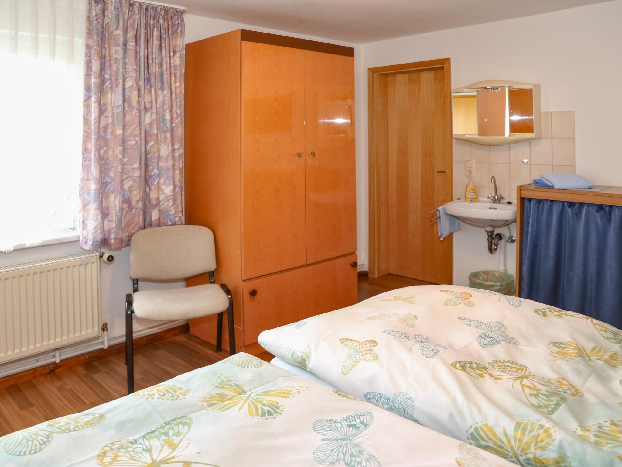 Ein Zimmer mit Bett, Stuhl, Kleiderschrank, einem Badezimmer mit Waschbecken und Spiegel sowie einem Fenster mit Vorhängen.