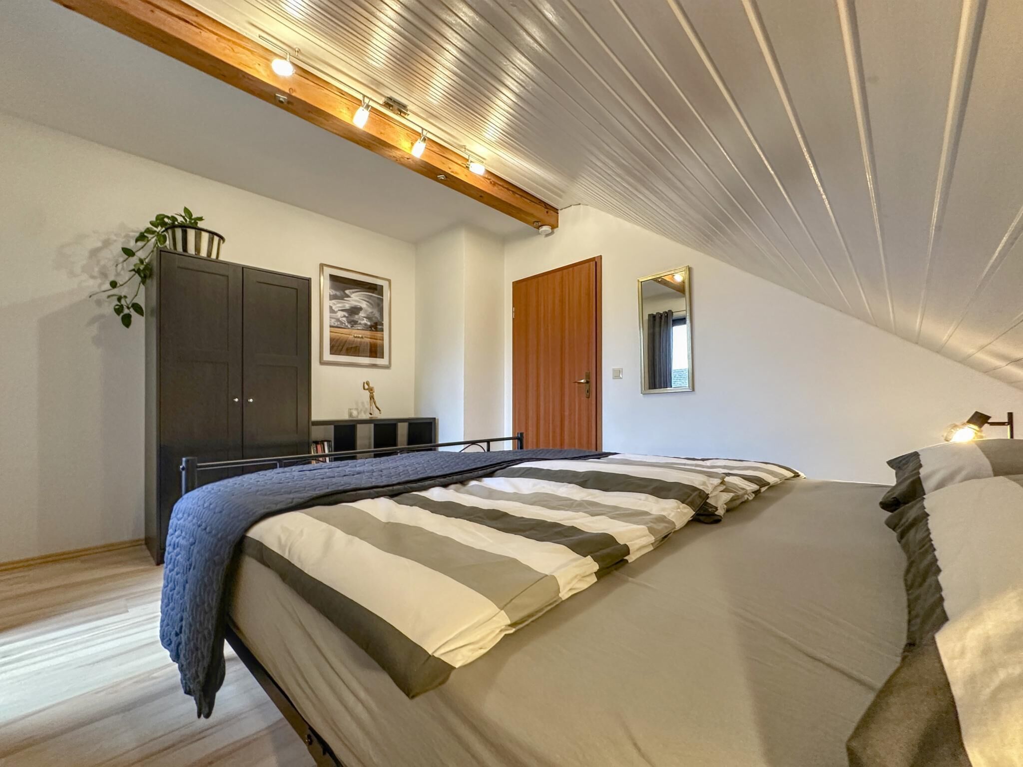 A bedroom with a large bed, a wardrobe, a mirror, and a ceiling with exposed beams and lights.