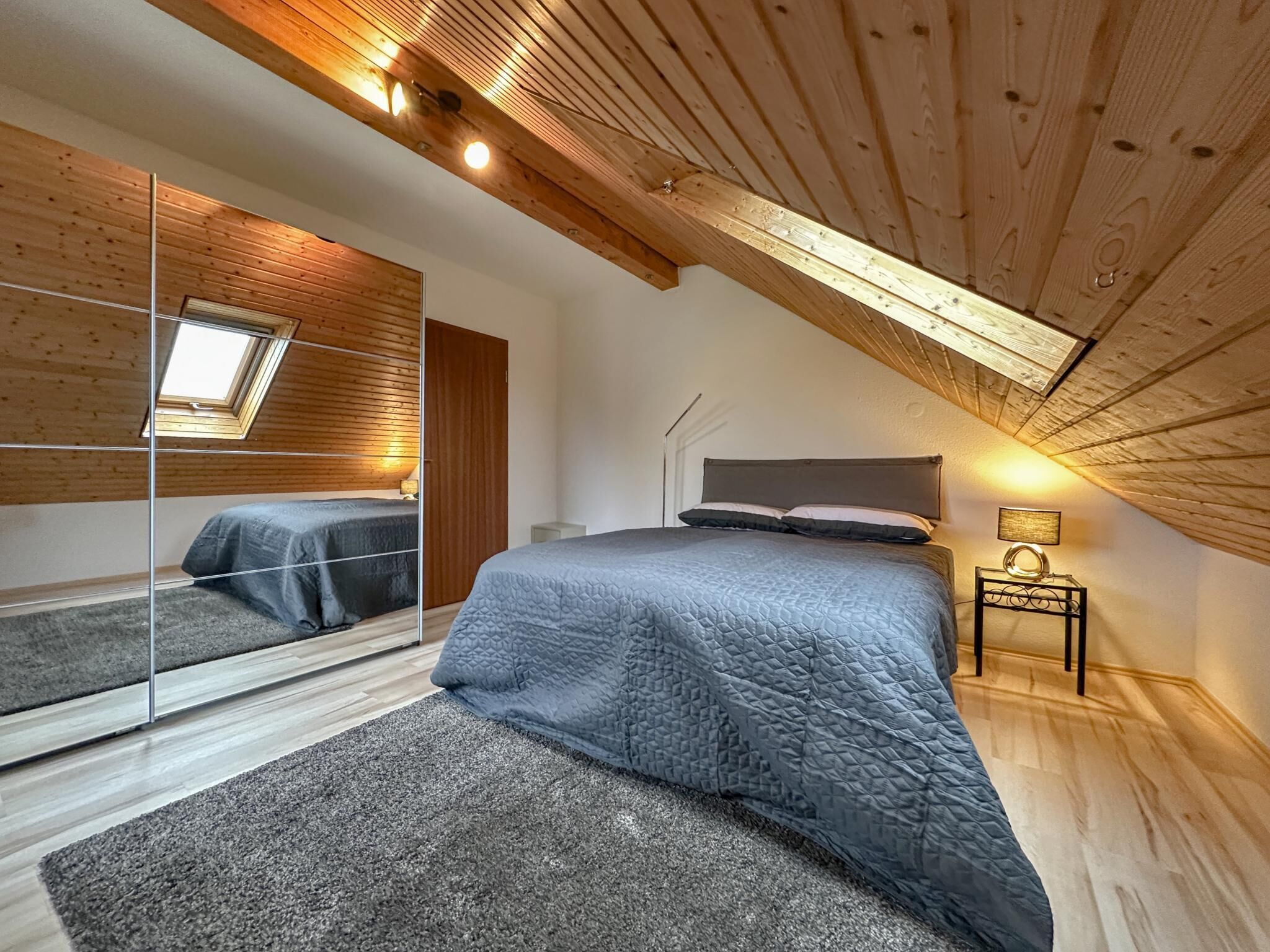 A bedroom with a sloped ceiling, wooden beams, a large bed with a quilted blanket, a bedside table with a lamp, and a skylight.