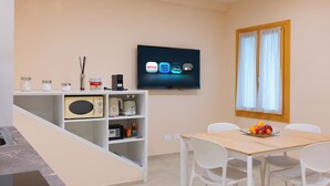 Eine moderne Küche mit Mikrowelle, Toaster und Kaffeemaschine auf einem weißen Regal, einem Esstisch mit Stühlen und einem Fernseher, der Streaming-Dienste anzeigt.