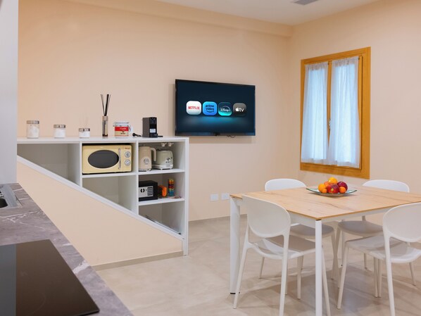 Eine moderne Küche mit Mikrowelle, Toaster und Kaffeemaschine auf einem weißen Regal, einem Esstisch mit Stühlen und einem Fernseher, der Streaming-Dienste anzeigt.
