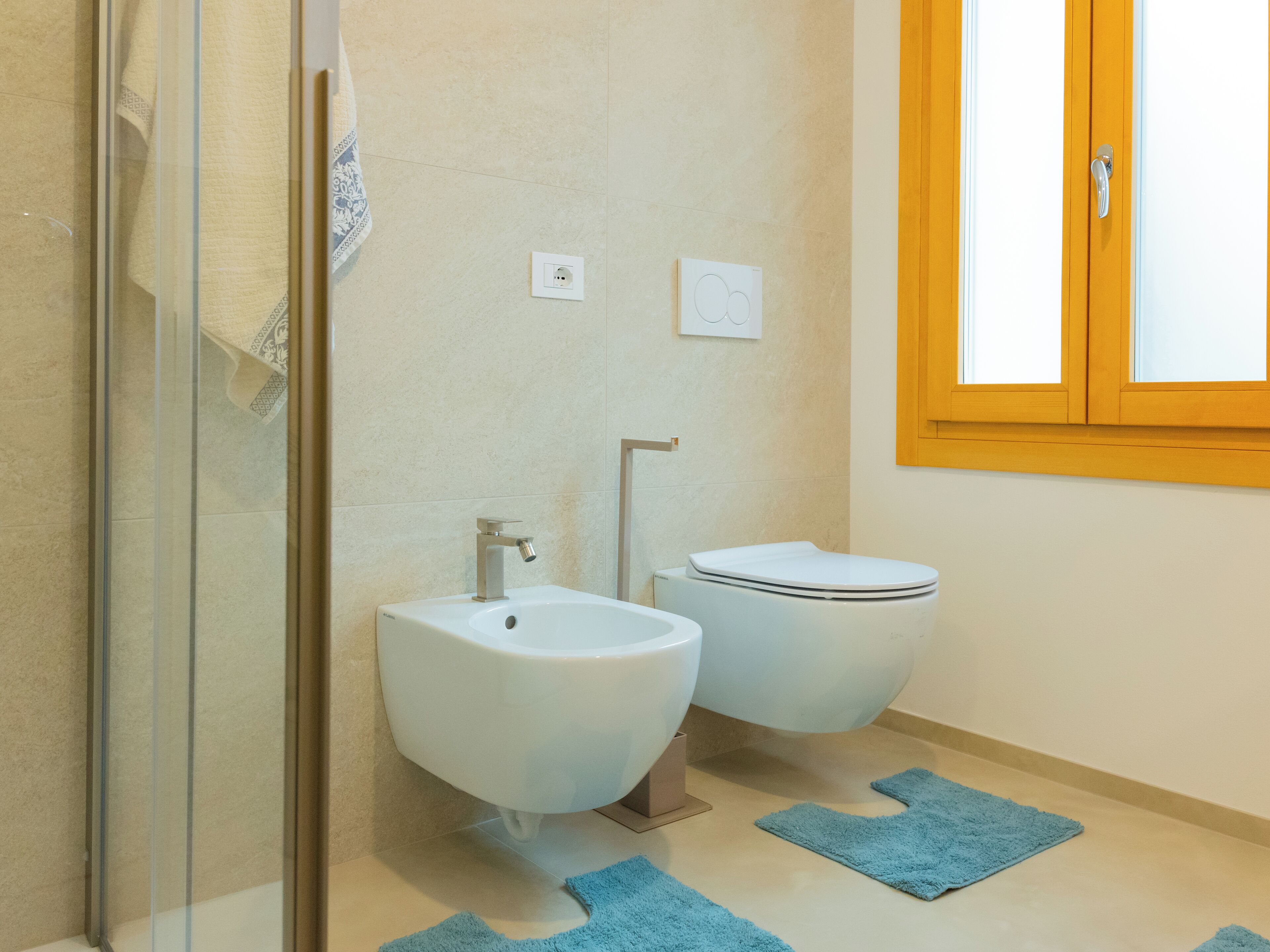 A bathroom with a glass shower enclosure, a bidet, a toilet, and a window with a wooden frame.