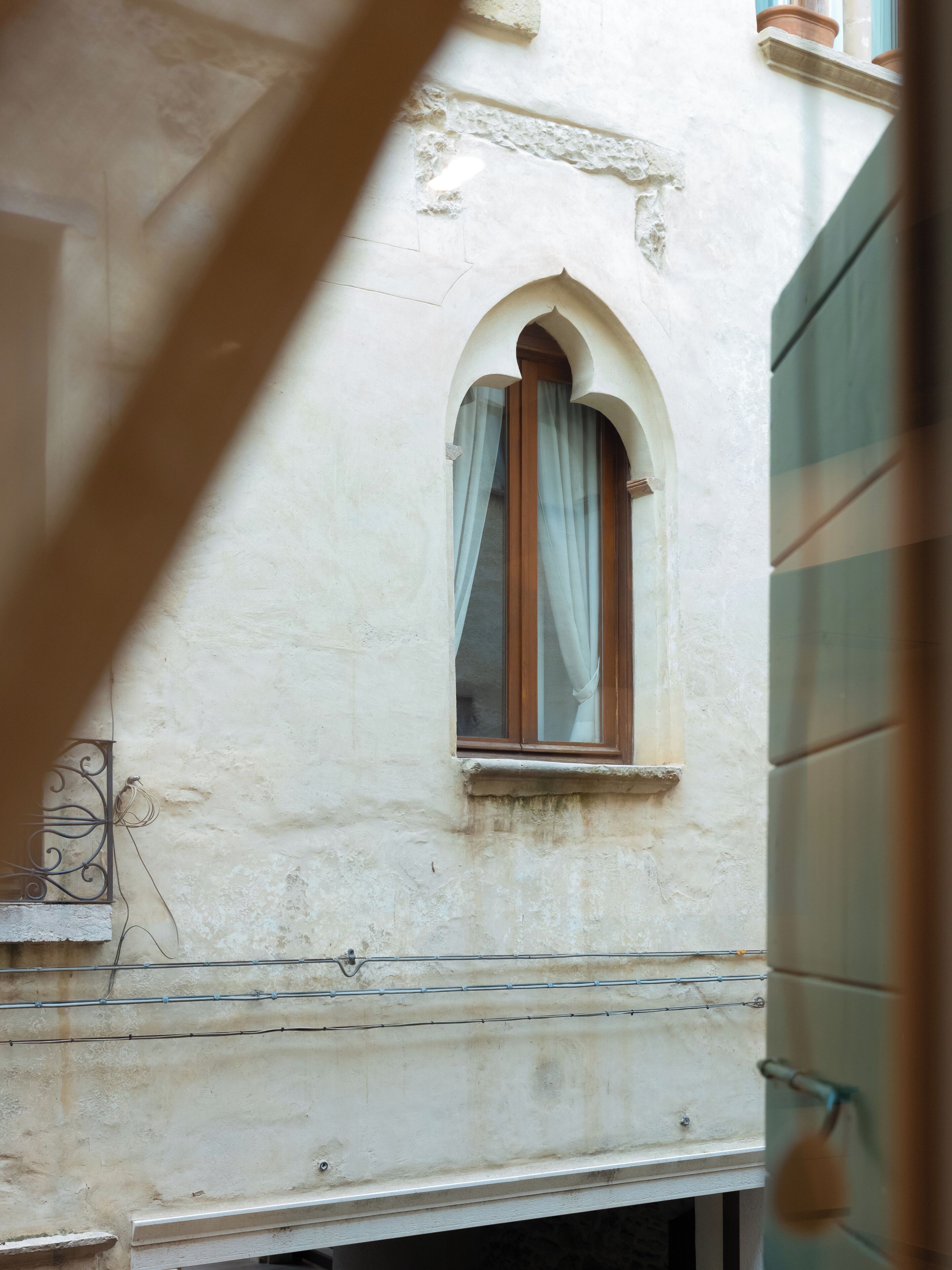 A narrow alleyway with a small arched window with a wooden frame and a white curtain.