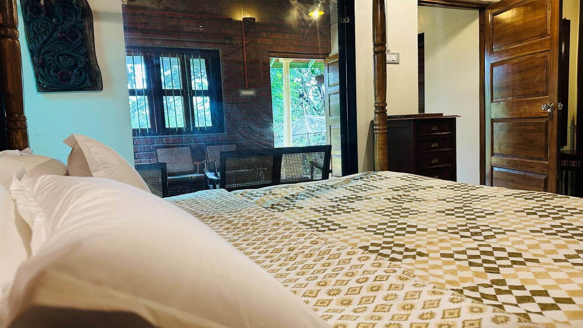Une chambre avec une couvre-lit à carreaux, une tête de lit en bois, une fenêtre avec des barreaux et une vue sur l’extérieur.