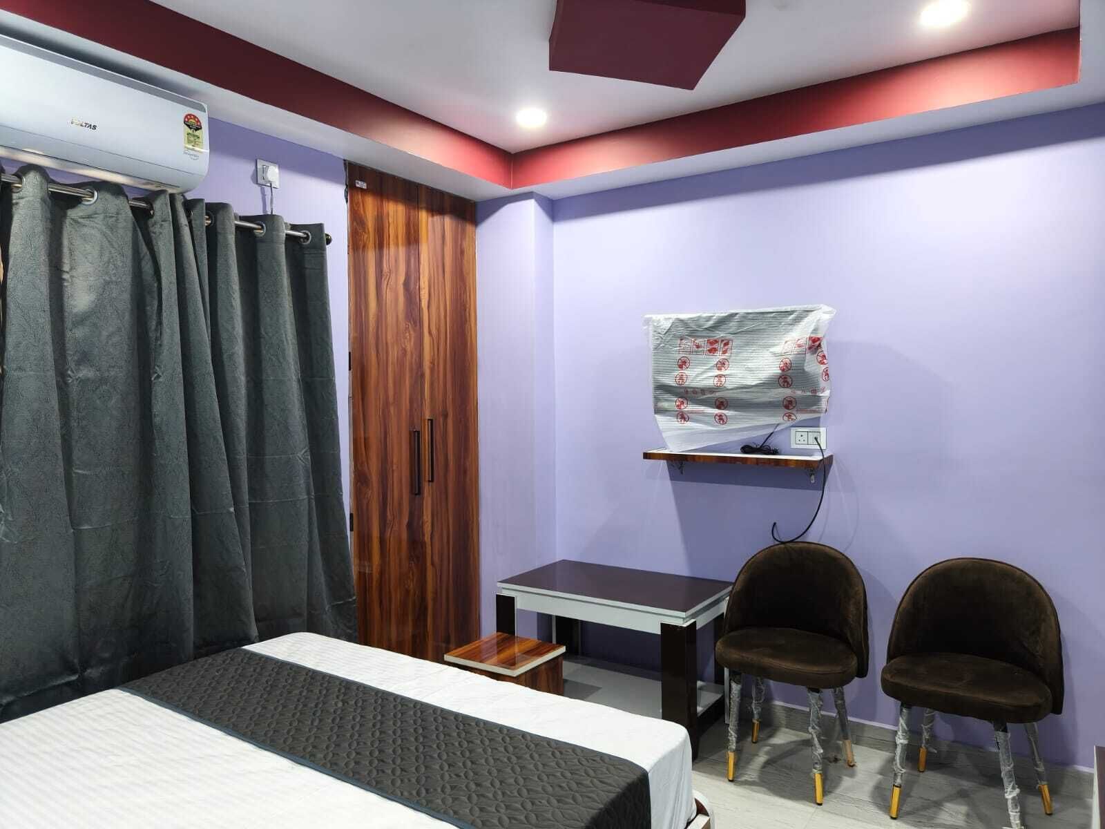 A bedroom with a bed, a desk, two chairs, a curtain, a wooden wardrobe, and a wall-mounted air conditioner.