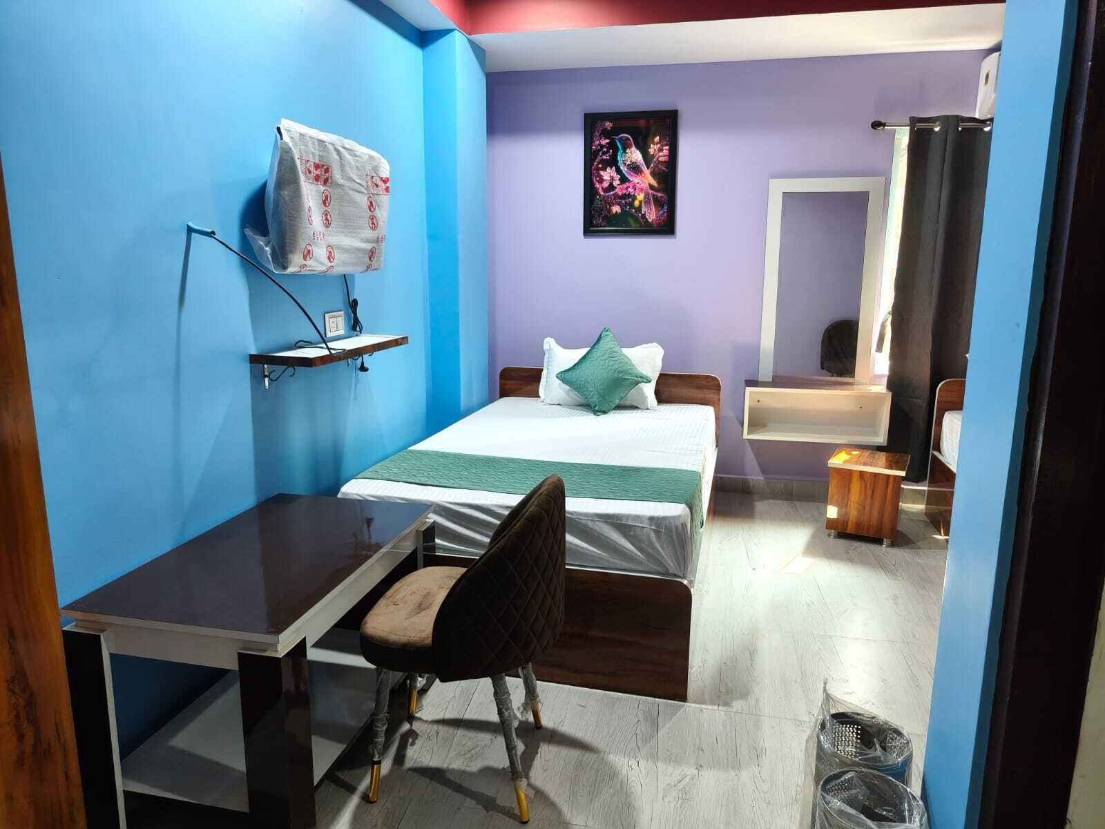 Une petite pièce, de taille modeste, comprenant un lit, un bureau et une chaise. On y trouve une étagère sur laquelle est posé un sac, un tableau encadré accroché au mur et un miroir.