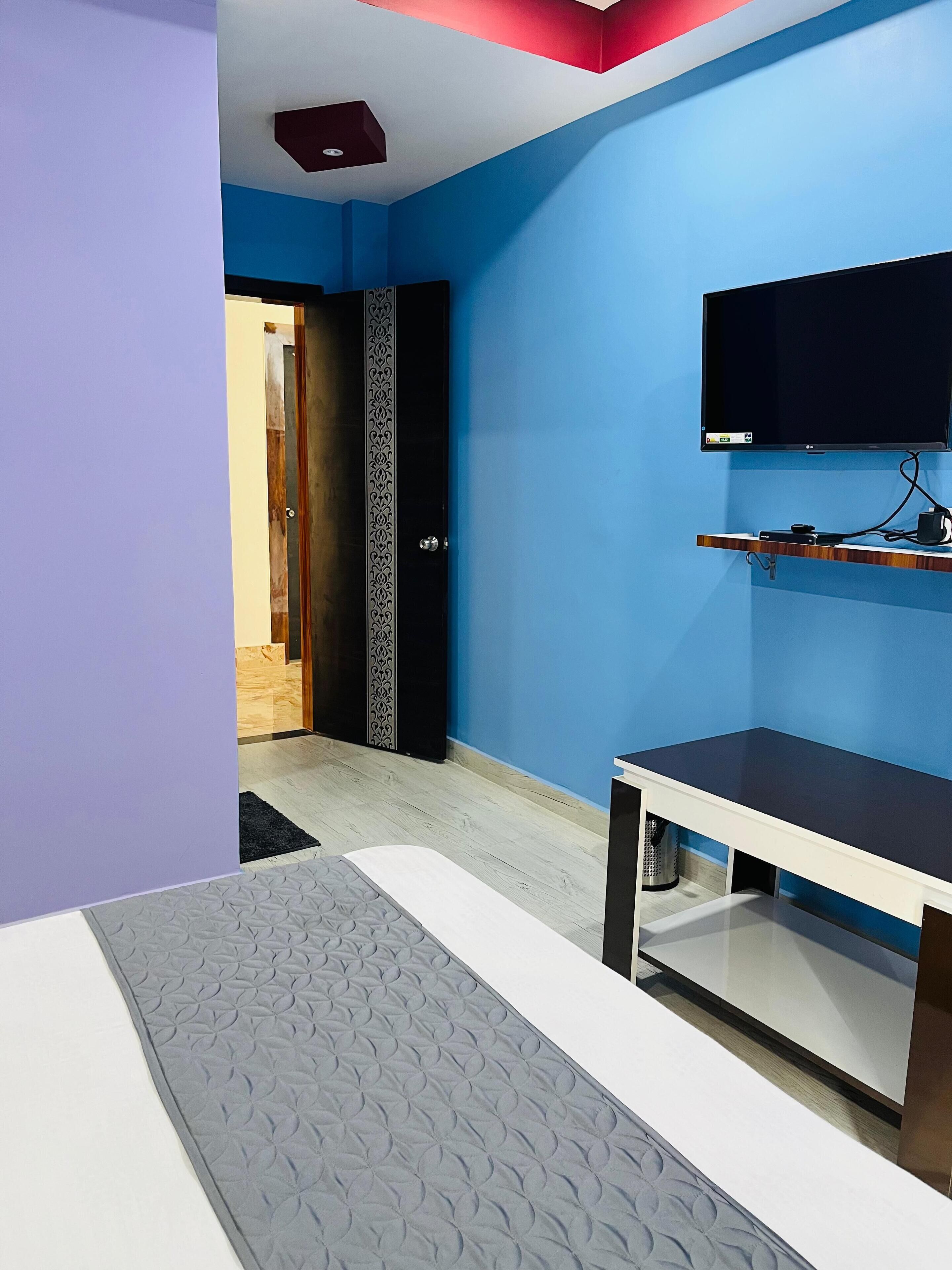 Habitación de hotel con pared azul, un televisor de pantalla plana montado sobre un estante de madera, cama con colcha gris estampada y una puerta que da a otra habitación.