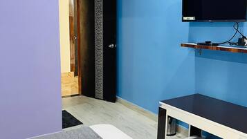 Habitación de hotel con pared azul, un televisor de pantalla plana montado sobre un estante de madera, cama con colcha gris estampada y una puerta que da a otra habitación.