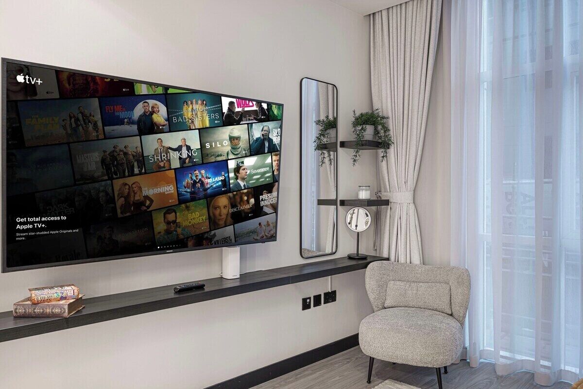 A modern living room with a large flat-screen TV mounted on the wall displaying various streaming content. There is a comfortable armchair and a small table with a decorative item on it.