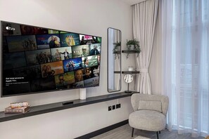 Ein modernes Wohnzimmer mit einem großen Flachbildfernseher an der Wand, der verschiedene Streaming-Inhalte zeigt. Ein bequemer Sessel und ein kleiner Tisch mit einem dekorativen Gegenstand stehen dort.