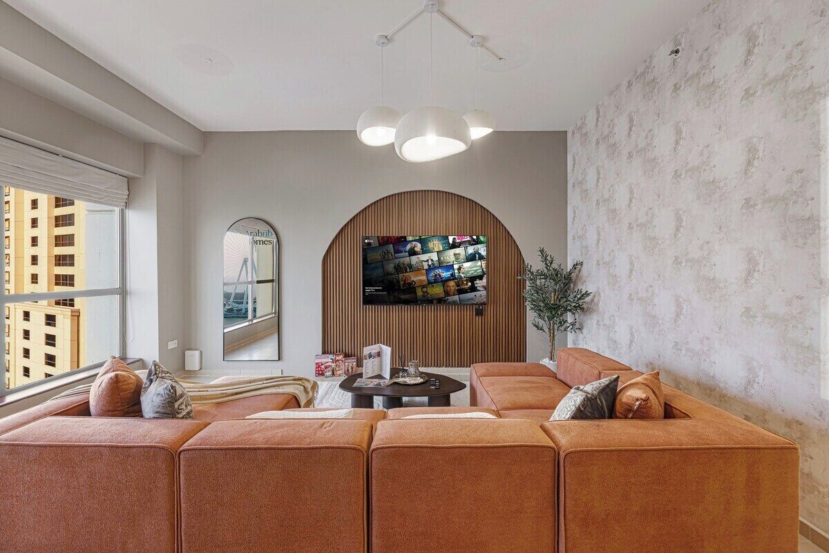 Un salon moderne avec un canapé d’angle, un téléviseur à écran plat fixé sur un panneau en bois et une grande fenêtre offrant une vue sur les bâtiments.