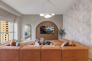 A modern living room with a sectional sofa, a flat-screen TV mounted on a wooden panel, and a large window providing a view of buildings. - JBR Walk / Ocean Views / Pool Table / Gym / Pool (Dubai)