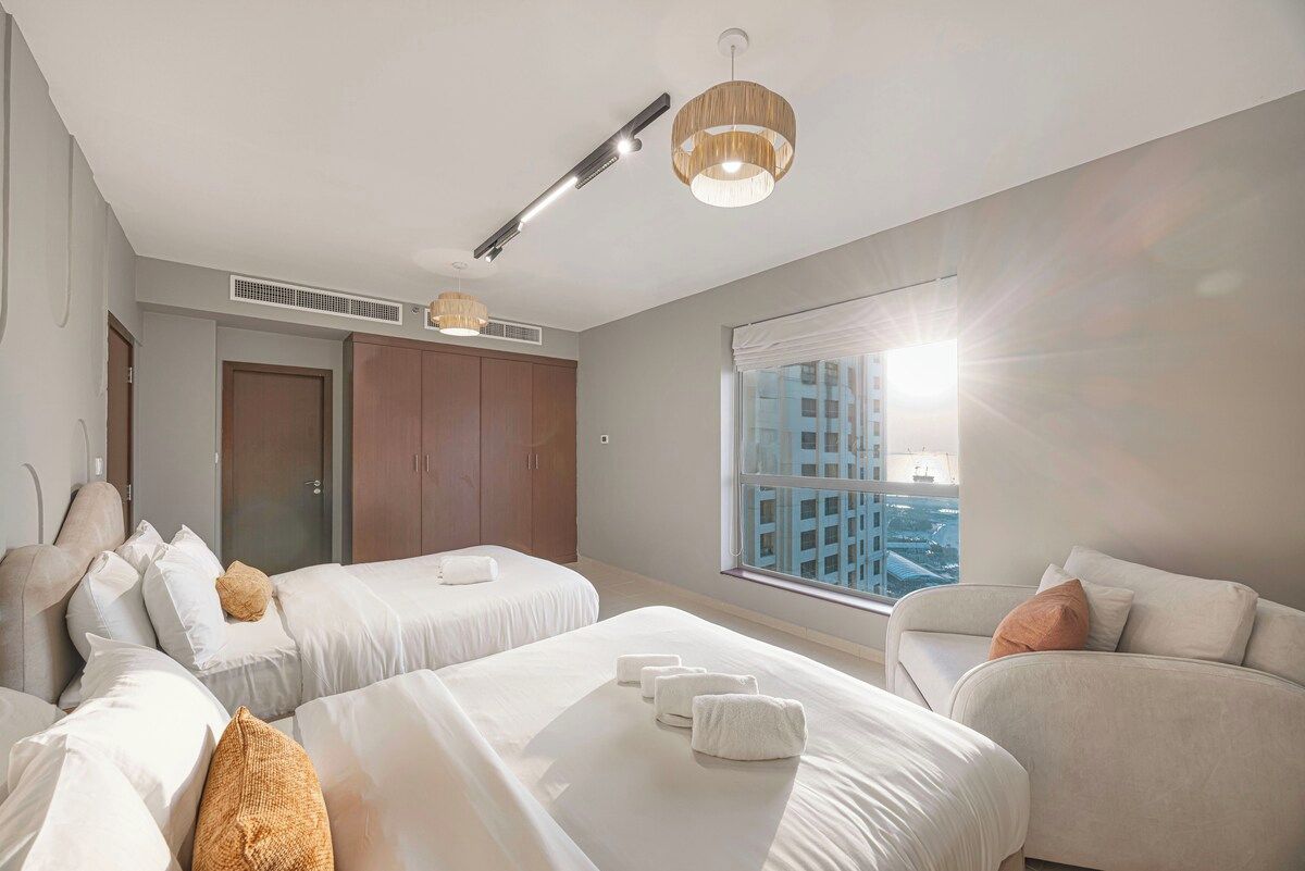 Ein Hotelzimmer mit zwei Betten, einer Couch, einem großen Fenster mit Blick auf die Stadt und einem Deckenventilator.