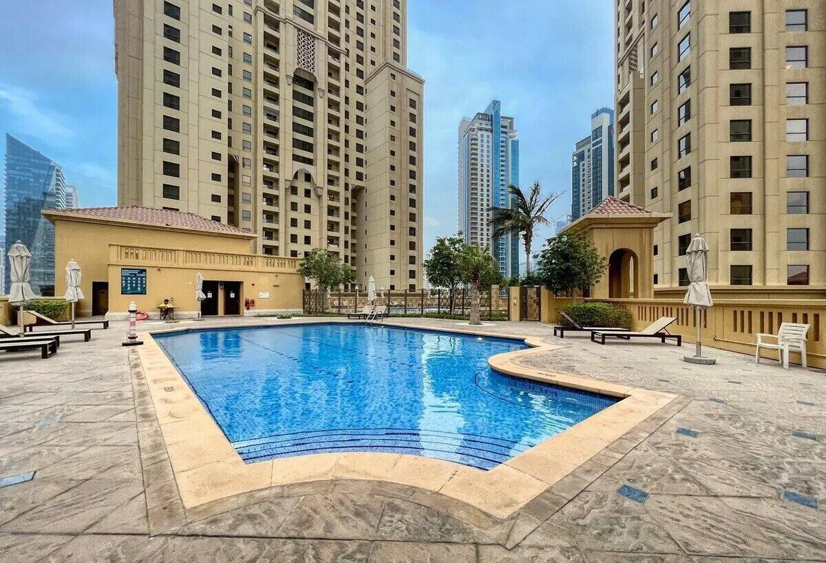 A swimming pool surrounded by high-rise buildings, with outdoor seating and a clear sky.