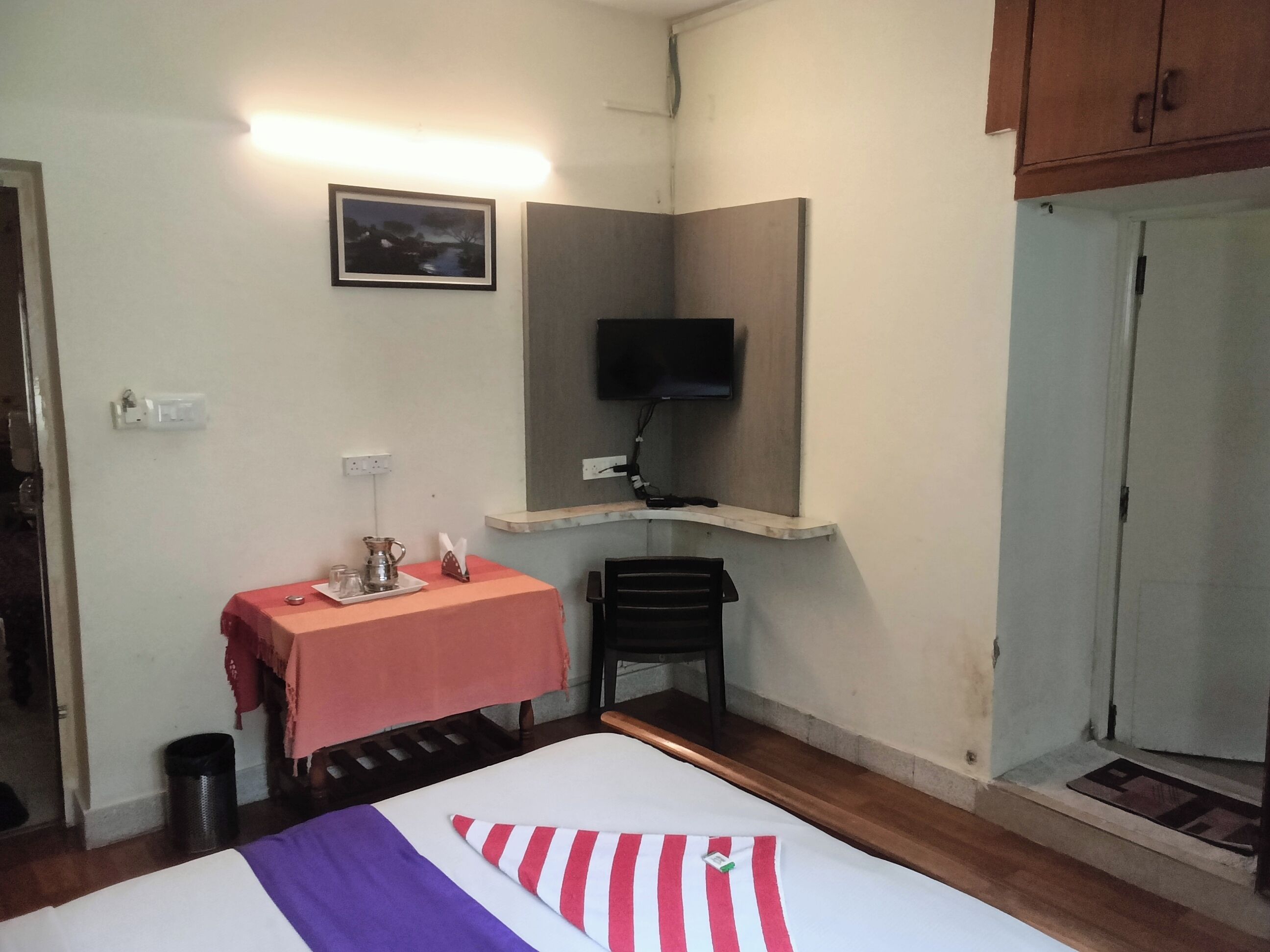 A hotel room with a bed, a table with a pitcher, a television mounted on a wall, and a door.