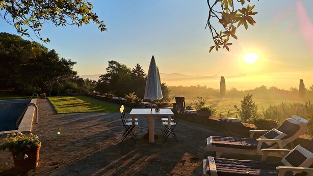 Um pátio externo com mesa e cadeiras, piscina e vista do pôr do sol sobre uma paisagem nevoenta.