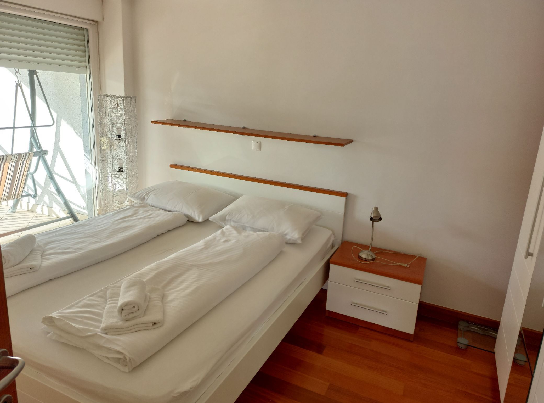 Ein Schlafzimmer mit Bett, Nachttisch, Lampe, einem Fenster mit Jalousien und einem Holzregal über dem Bett.