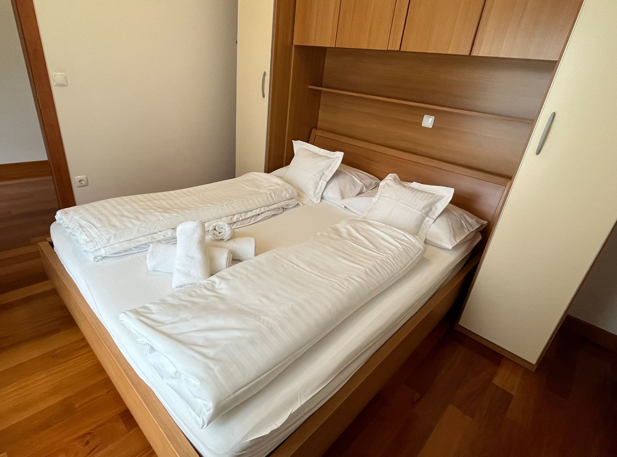 Un lit double avec du linge de lit blanc et deux serviettes enroulées, une tête de lit en bois et un plancher en bois.