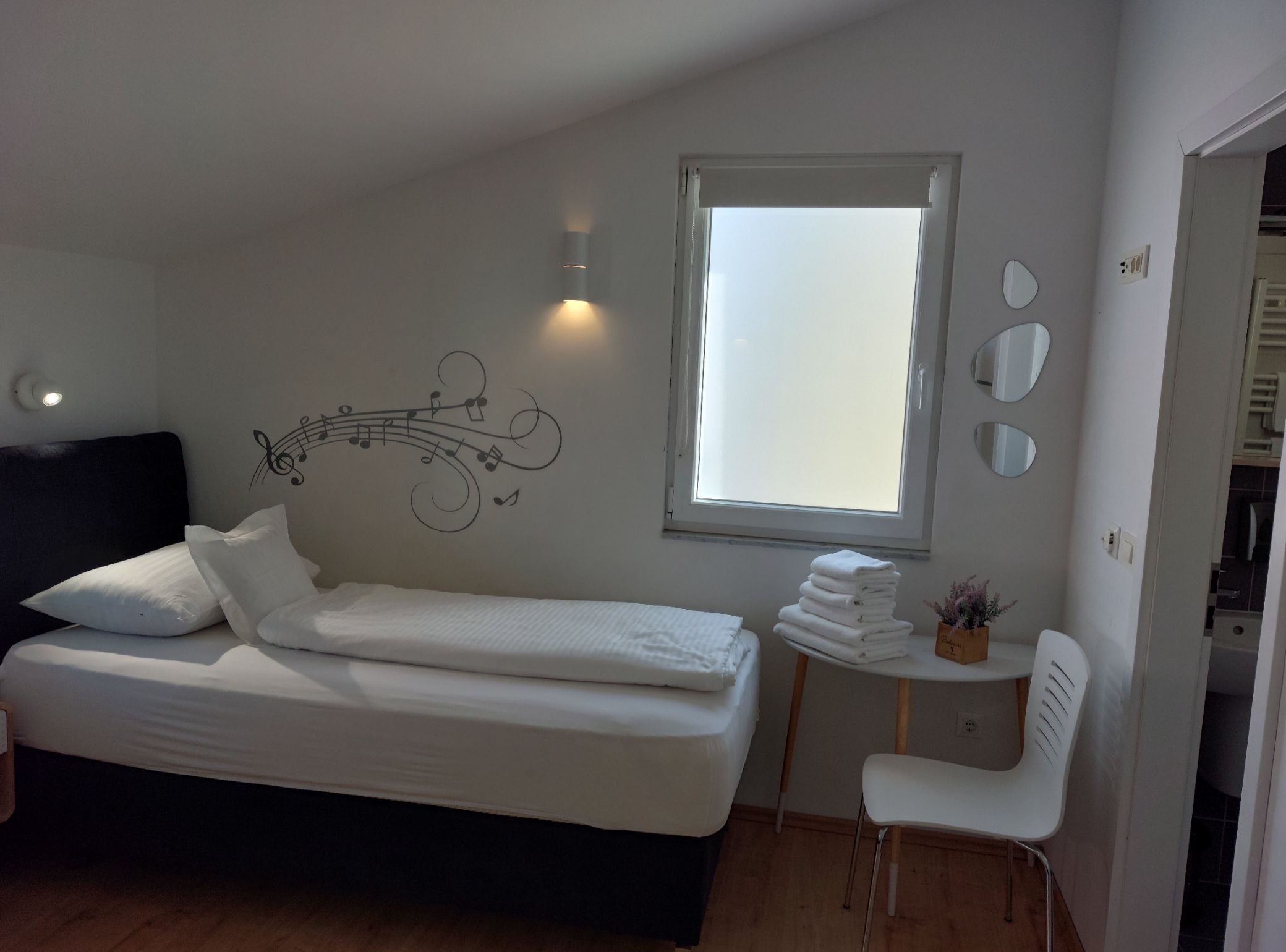 Una camera da letto con un letto, un tavolino con asciugamani, una sedia, una finestra e un addobbo a parete.