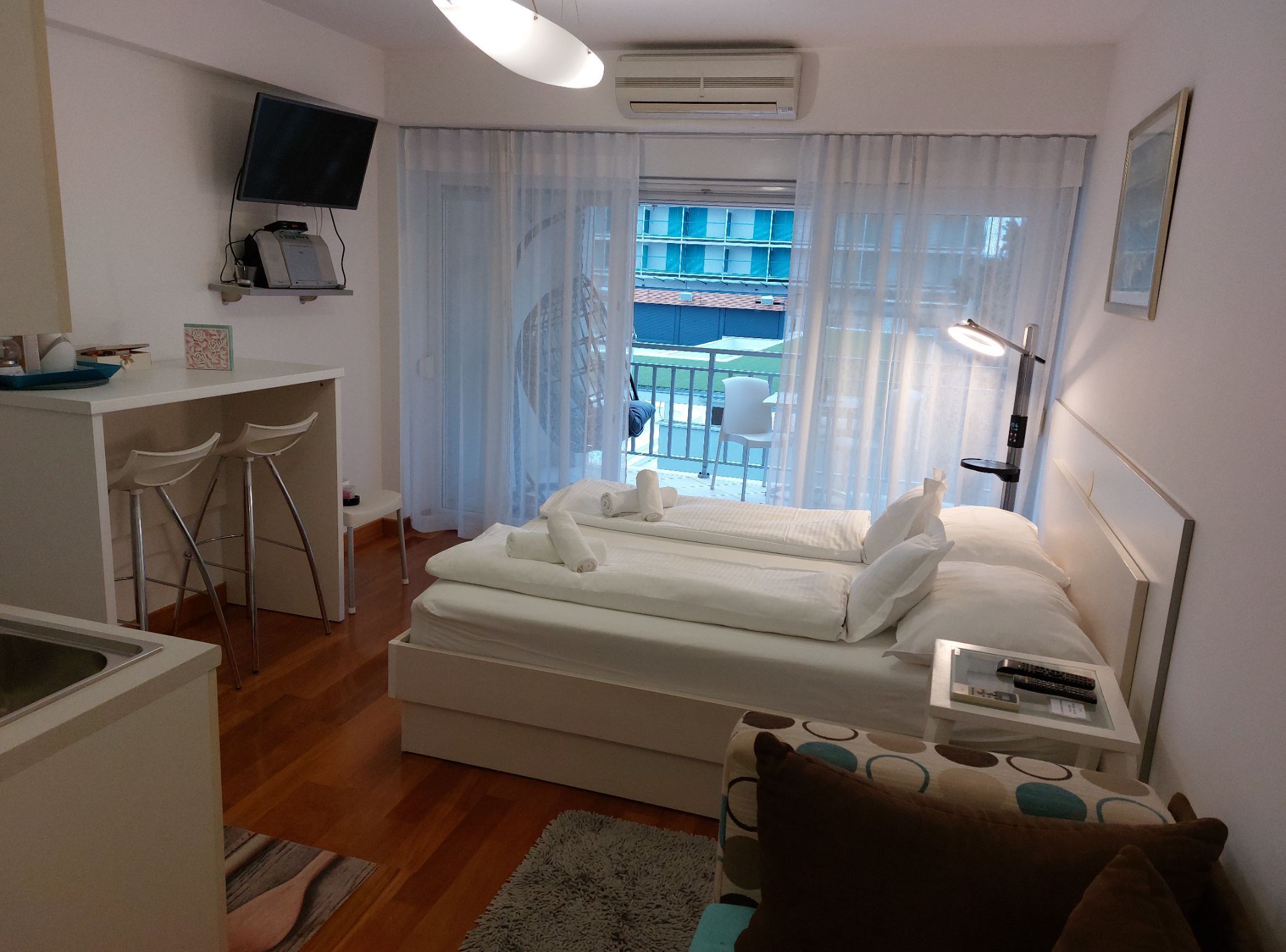 A hotel room with a bed, a desk with a chair, a TV, and a balcony with a view.