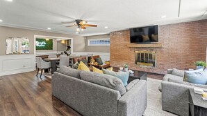 A spacious living room with a fireplace, grey sofas, and a dining area with a brick wall and ceiling fan. - Endless Family Entertainment, Fire Pit, 2 Kings! (Wichita)