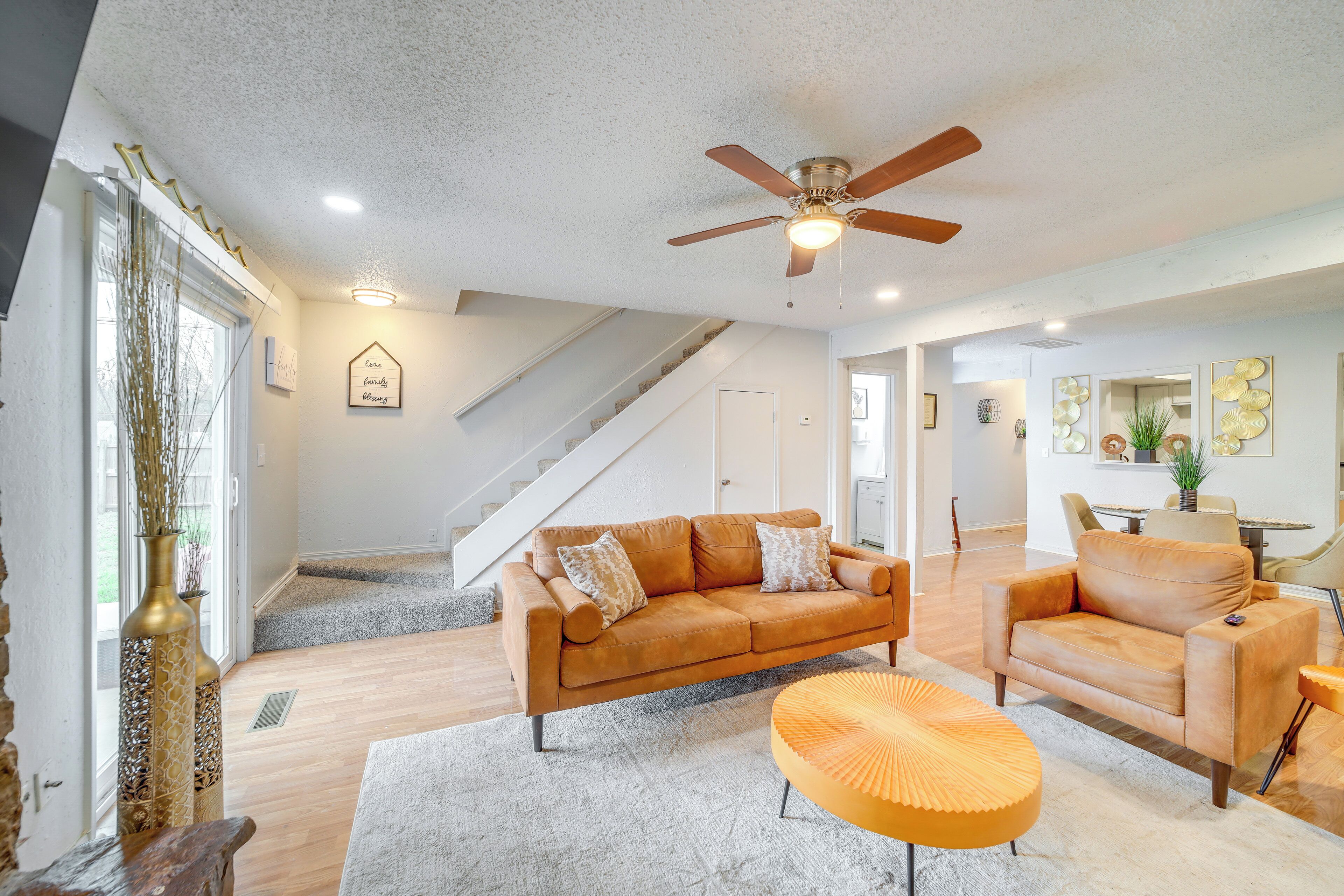 A spacious living room with a sofa, armchairs, and a coffee table. A ceiling fan and a staircase are visible in the background.