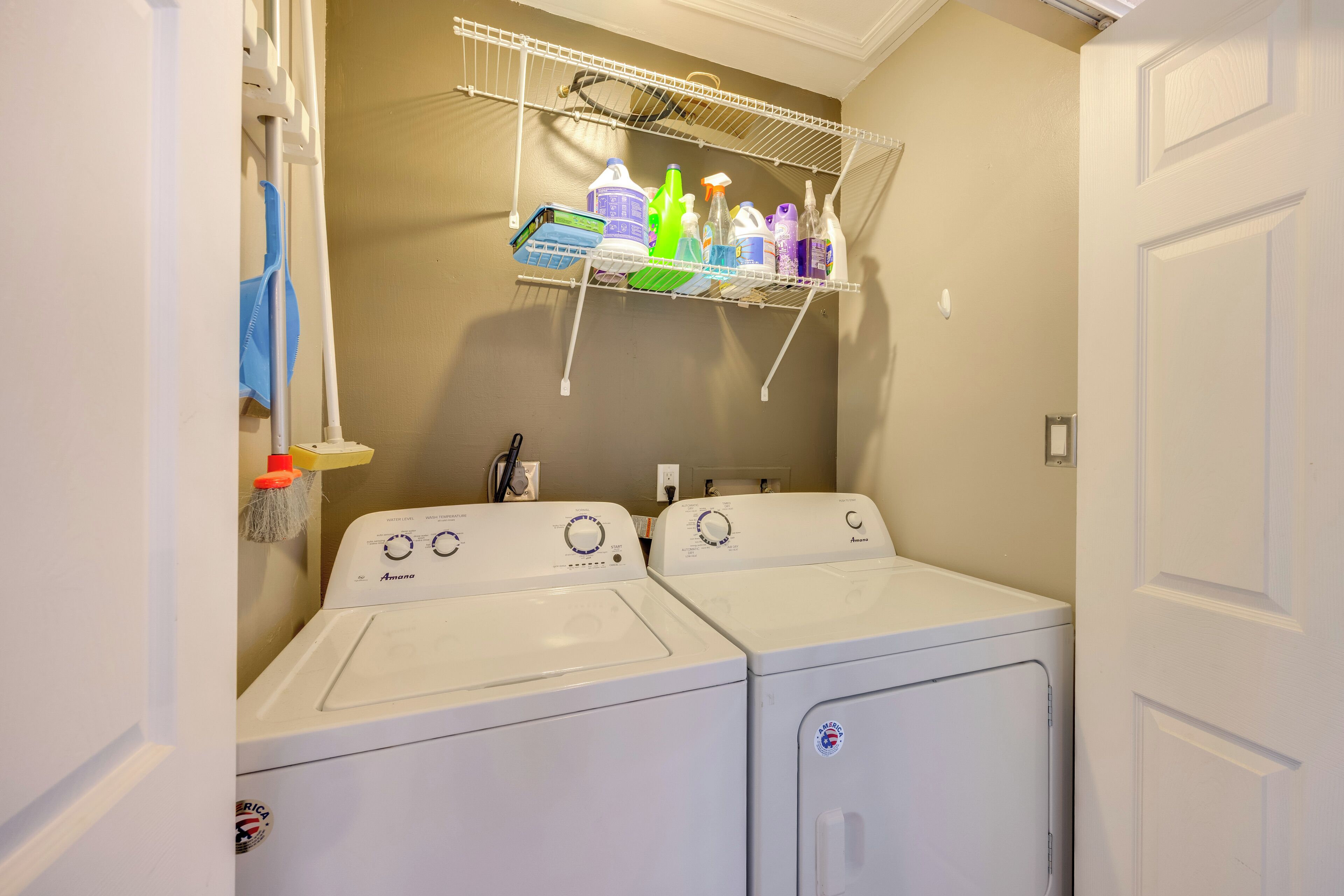 A laundry room with two front-loading washing machines, a shelf above holding cleaning supplies, and a door to the right.