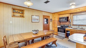 A wooden dining table with benches, a kitchen area with a microwave and stove, and a window with blinds.