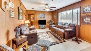 A cozy living room with wooden walls, a ceiling fan, a flat-screen TV, and a large window with blinds.