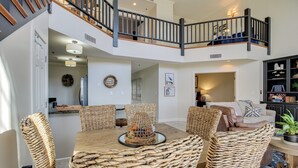 A dining area with a wooden table and woven chairs, a staircase with a railing, and a living room with a sofa and decorative items.
