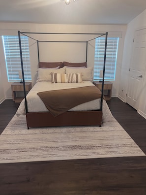 Ein Bett mit Baldachin, ein braunes Kopfende, eine weiße Bettdecke und mehrere Kissen. Neben dem Bett steht ein Nachttisch mit einer Lampe. Der Raum hat dunkle Holzböden und einen gemusterten Teppich.