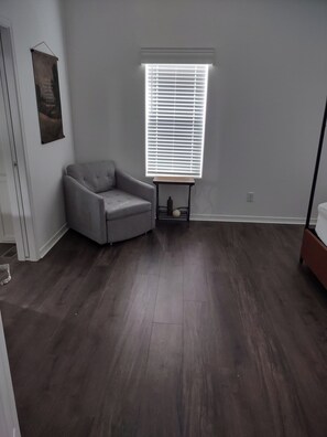 Ein Zimmer mit grauem Sessel, einem Fenster mit Jalousien und einem kleinen Tisch mit einer Flasche.
