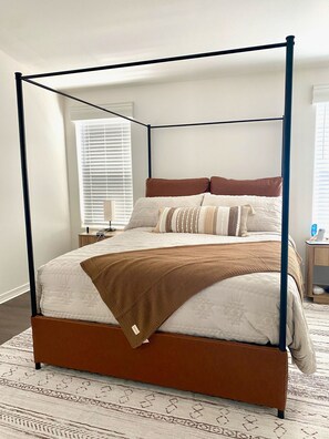 Ein Bett mit Baldachin, dessen Gestell braun, die Bettwäsche weiß und die Tagesdecke gemustert ist.