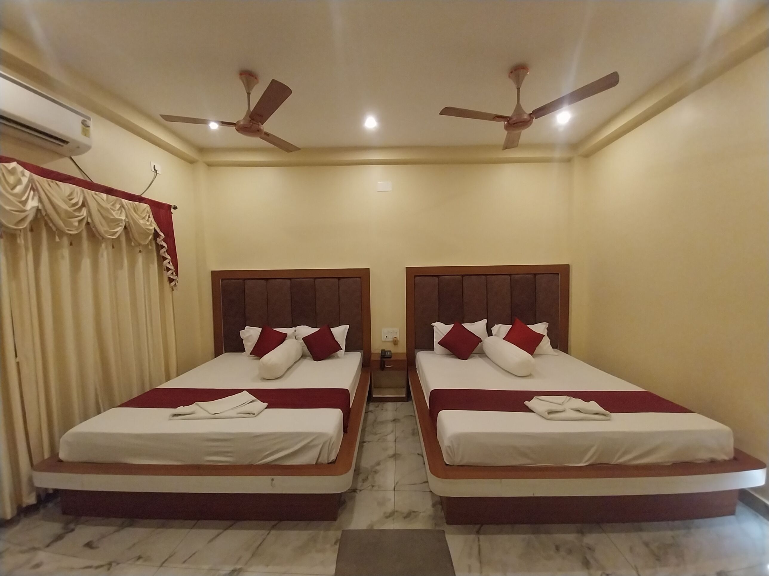 A hotel room with two beds, each with a wooden headboard and white bedding. There are ceiling fans and an air conditioning unit.