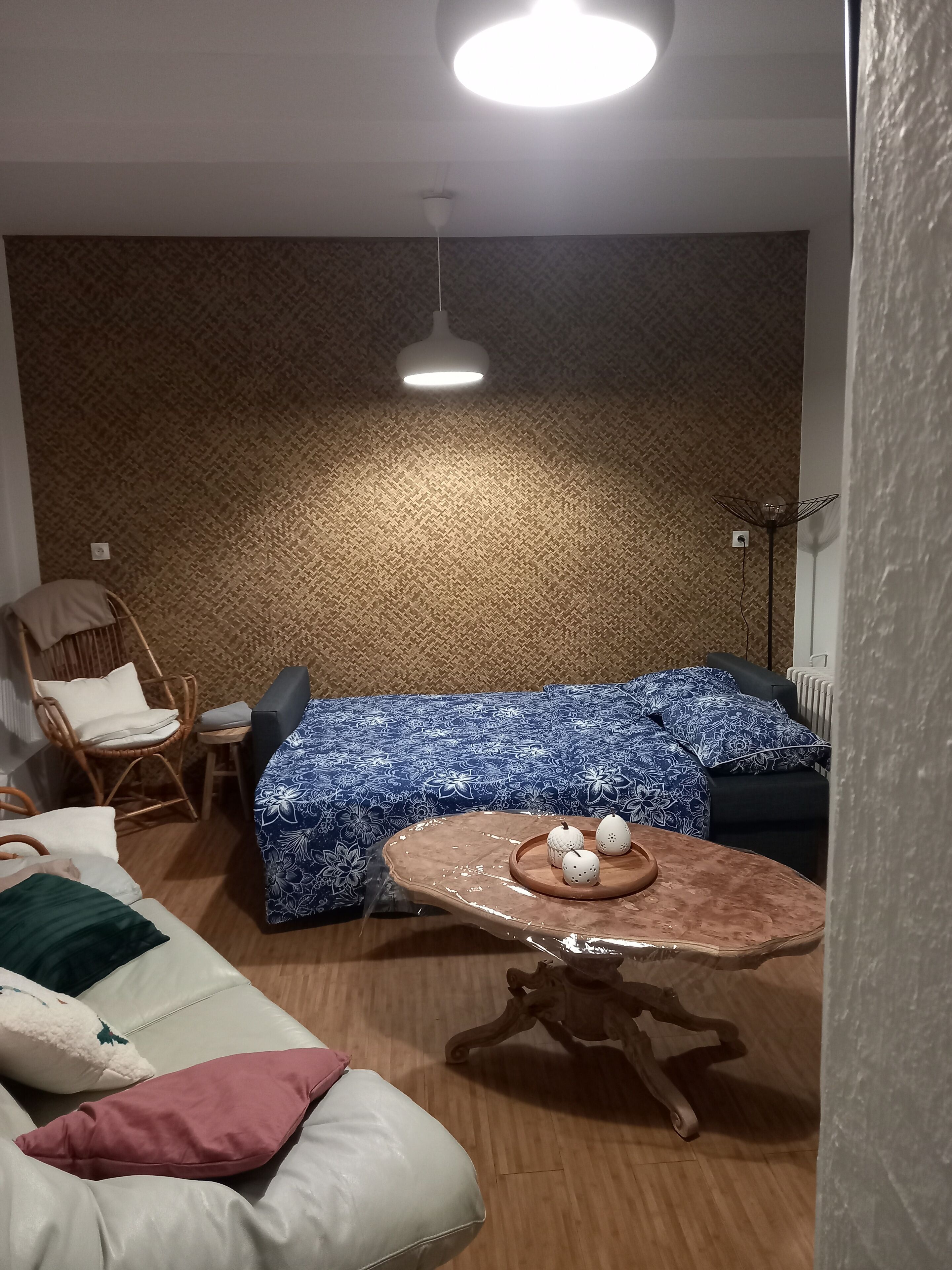 A cozy room with a patterned wall, a bed with a blue floral bedspread, a wicker chair, a small table with decorative items, and a floor lamp.