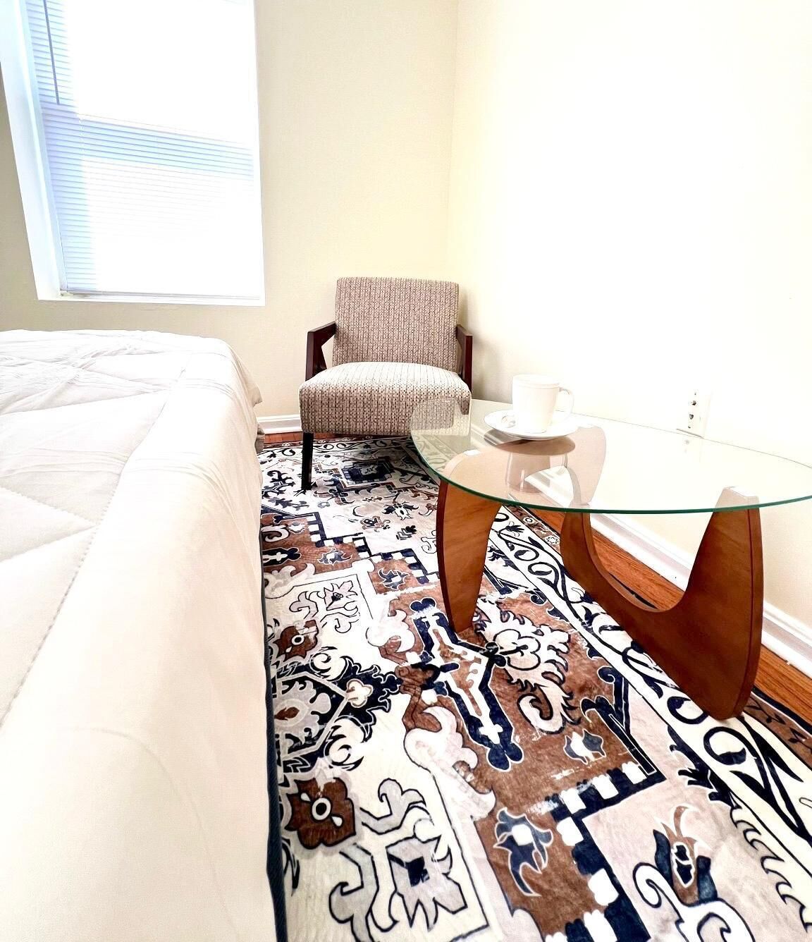 A hotel room with a bed, a patterned rug, a chair, a glass coffee table, and a window.