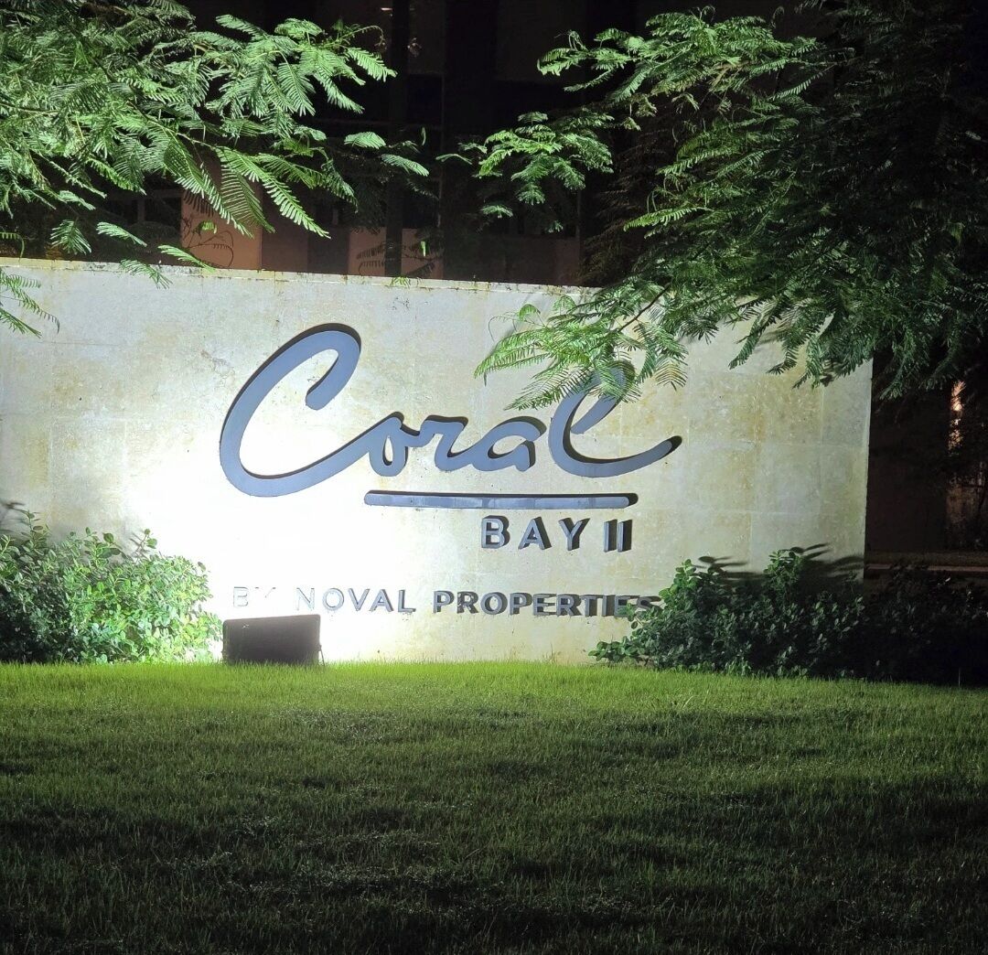 A sign for Coral Bay II, a property by NOVAL PROPERTIES, with a grassy foreground and trees in the background.