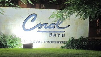 A sign for Coral Bay II, a property by NOVAL PROPERTIES, with a grassy foreground and trees in the background.