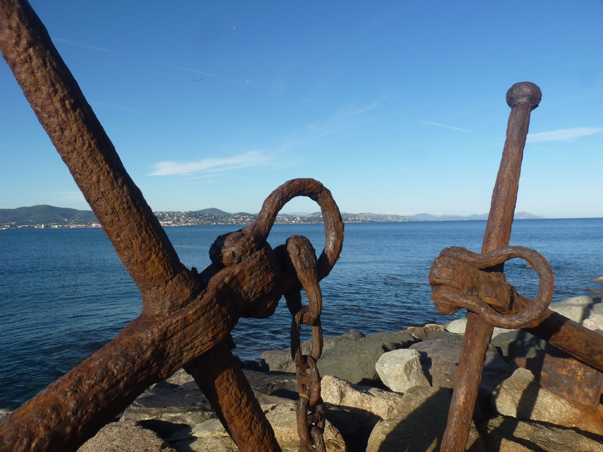 A rusty anchor and chain by the sea with a clear view of the water and distant land.