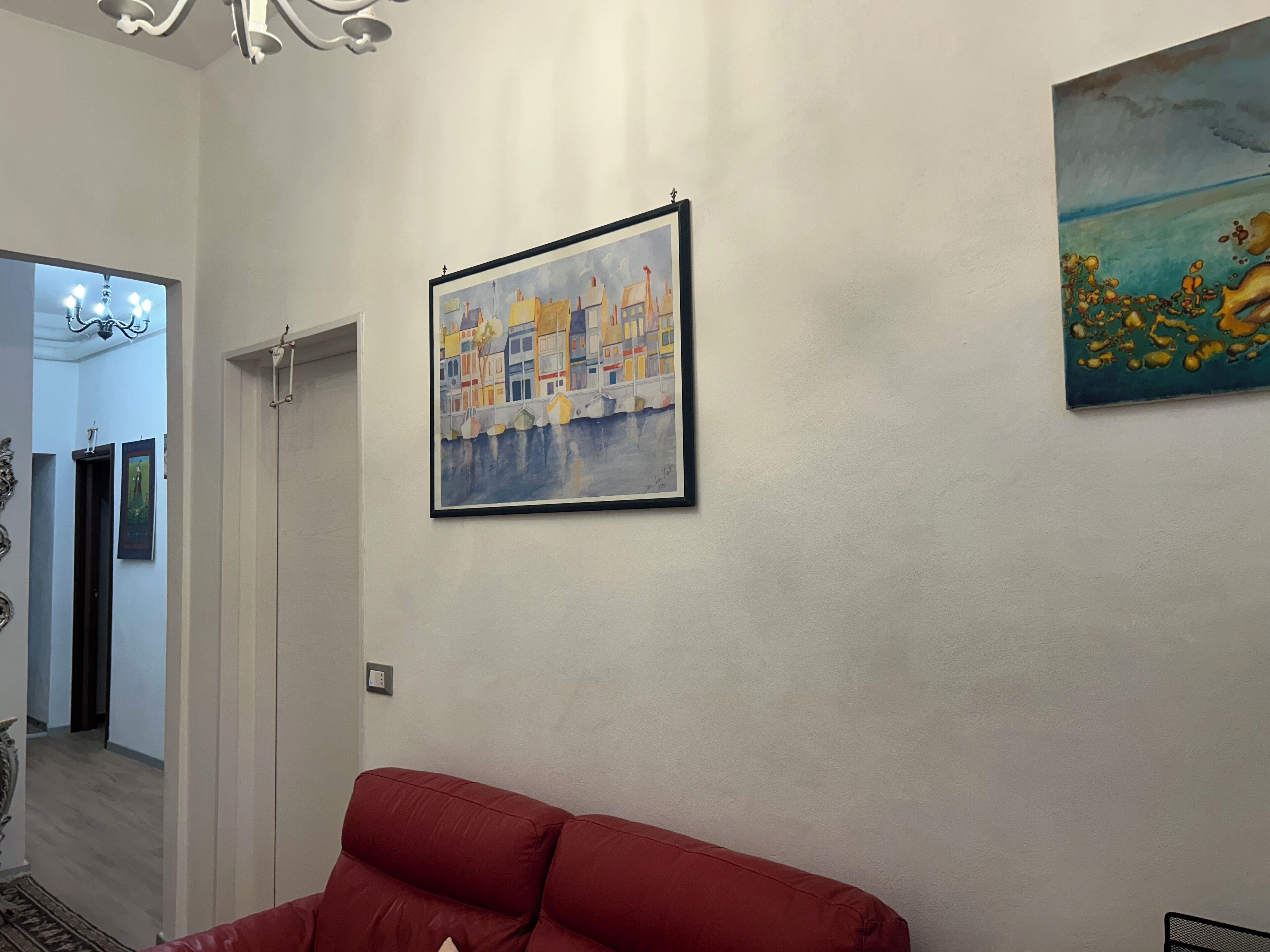 Una stanza con un divano rosso, un quadro incorniciato di un vivace paesaggio urbano e un lampadario.