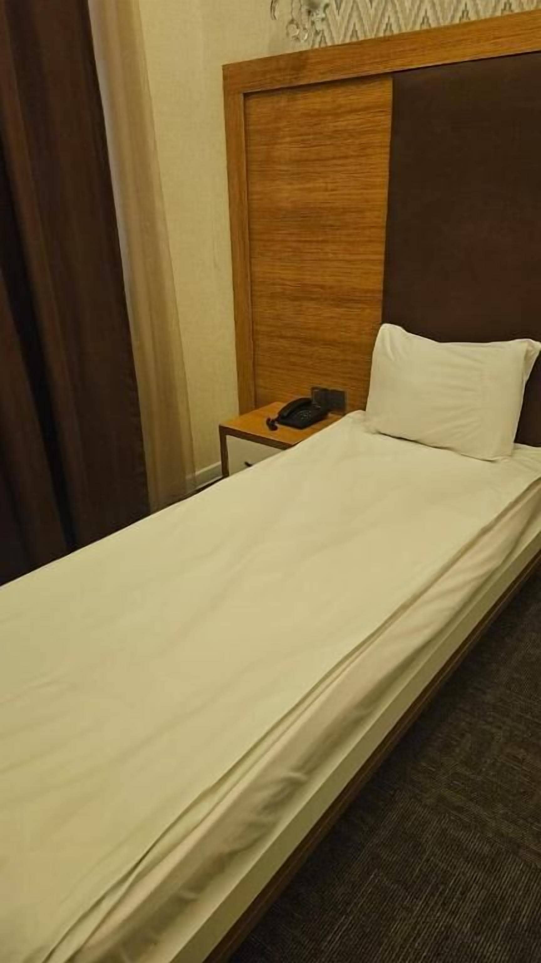 Ein Einzelbett mit Holz-Kopfteil, ein Nachttisch mit Telefon und ein Vorhang.