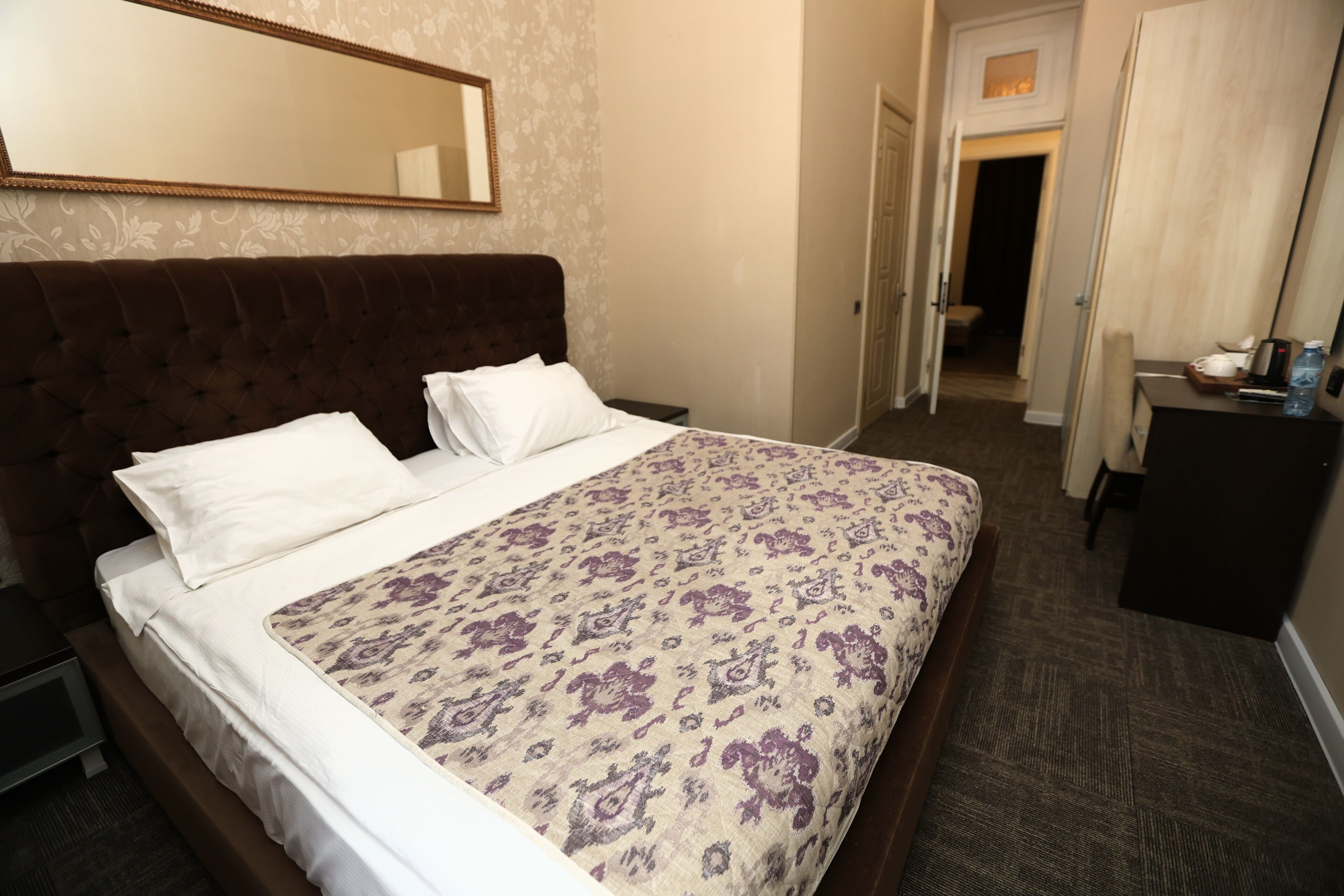 A hotel room with a bed, a bedside table, a dresser, a mirror, and a door leading to another room.
