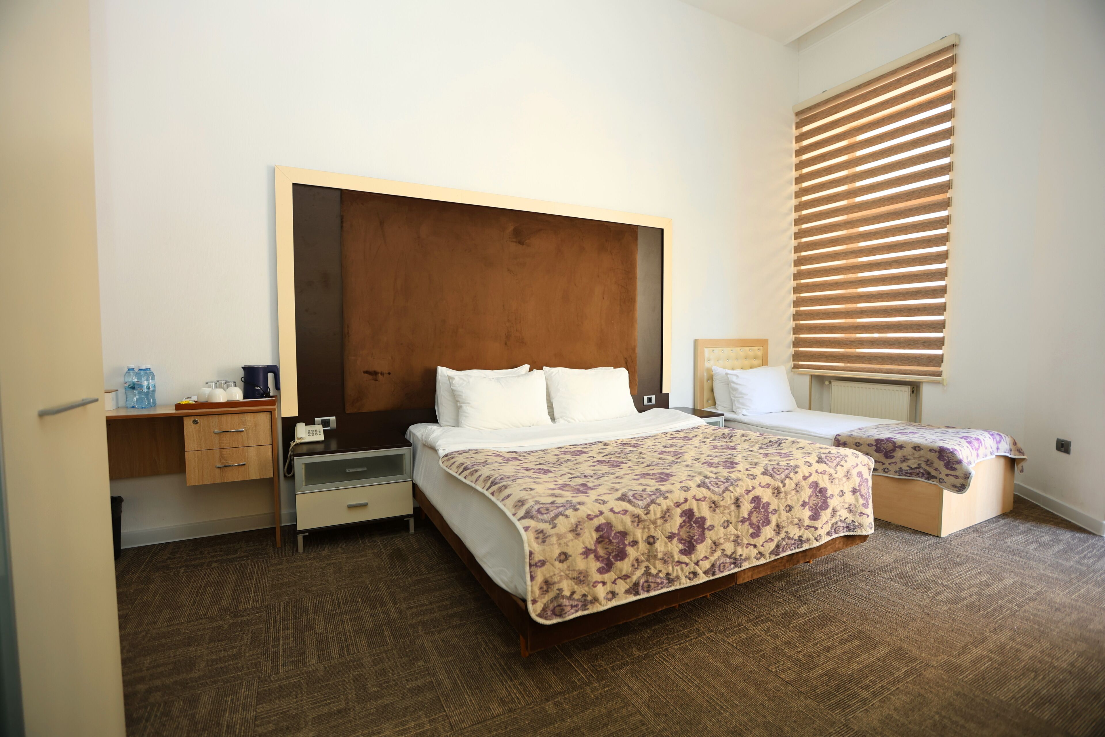 A hotel room with a large bed, a bedside table, a desk, and a window with blinds.