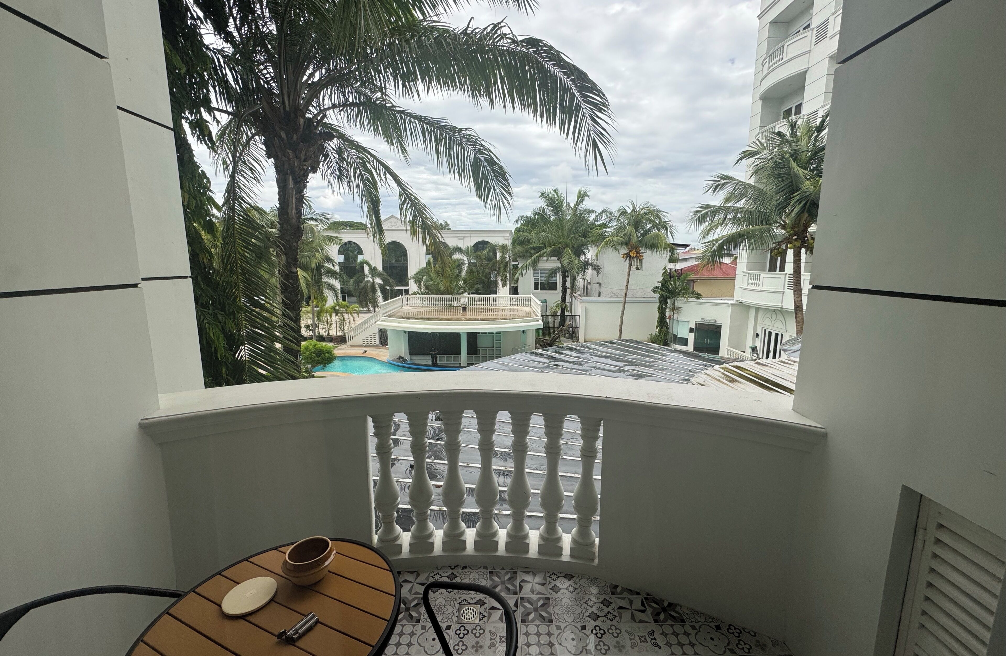 A balcony with a view of a pool, palm trees, and a building with arched windows.