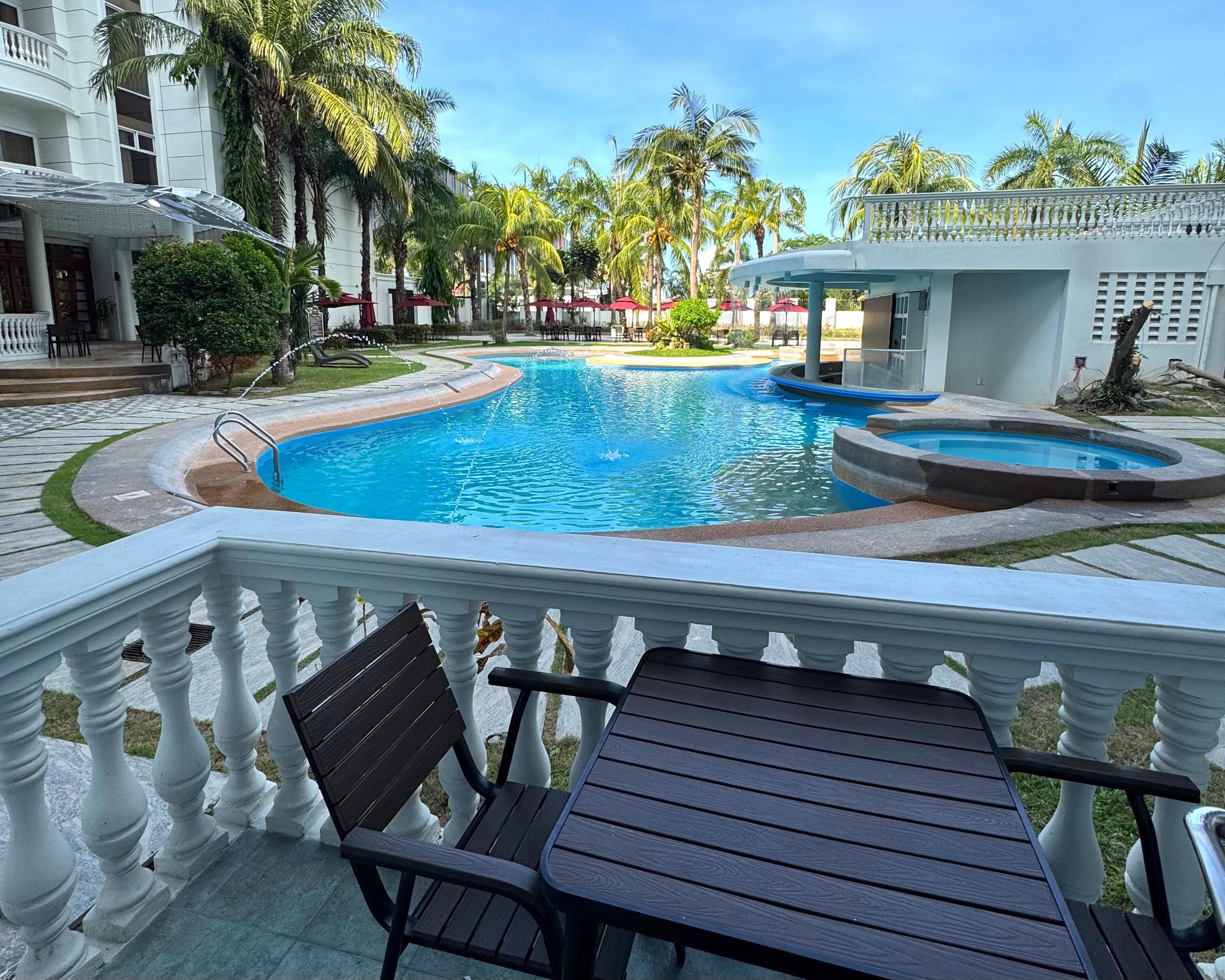 A swimming pool with a fountain, surrounded by palm trees and a white building with balconies.